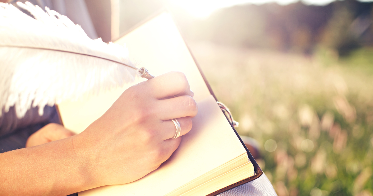 Writer’s journal in sunlight on grass with a woman’s hand holding a quill—capturing writing goals for May and creative freedom outdoors.