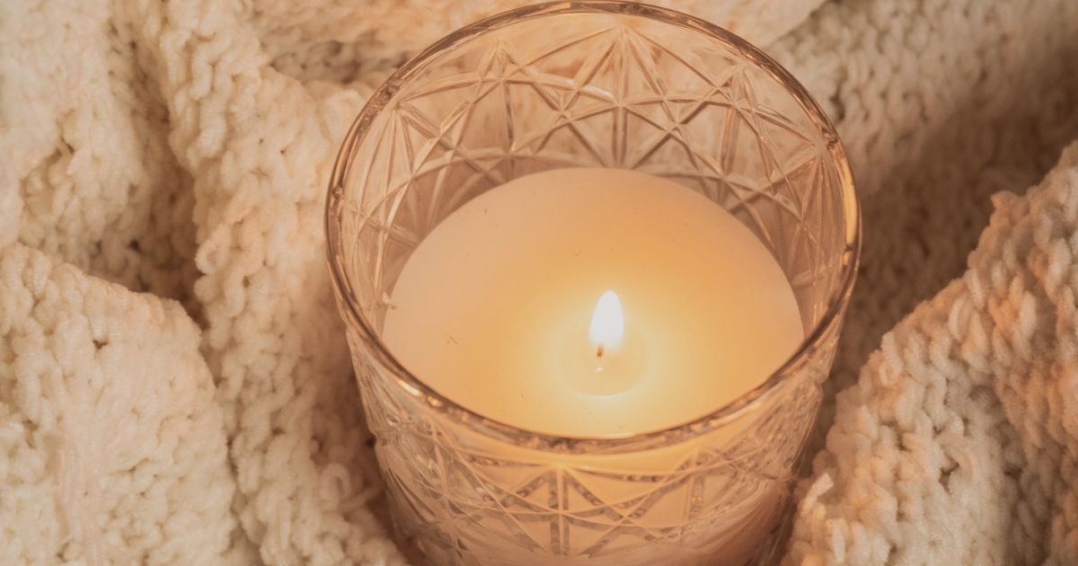 A lit candle in a cut-glass holder nestled in a soft cream knit blanket.